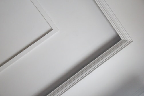 A close-up view of an interior ceiling corner with decorative molding. The light gray color of the wall contrasts slightly with the white trim, creating a subtle geometric pattern.