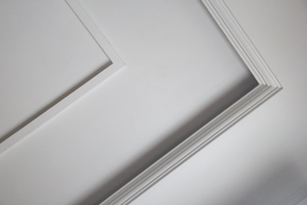 A close-up view of an interior ceiling corner with decorative molding. The light gray color of the wall contrasts slightly with the white trim, creating a subtle geometric pattern.