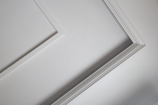A close-up view of an interior ceiling corner with decorative molding. The light gray color of the wall contrasts slightly with the white trim, creating a subtle geometric pattern.