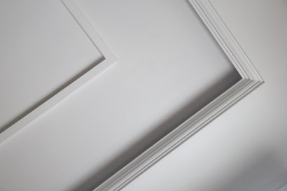 A close-up view of an interior ceiling corner with decorative molding. The light gray color of the wall contrasts slightly with the white trim, creating a subtle geometric pattern.