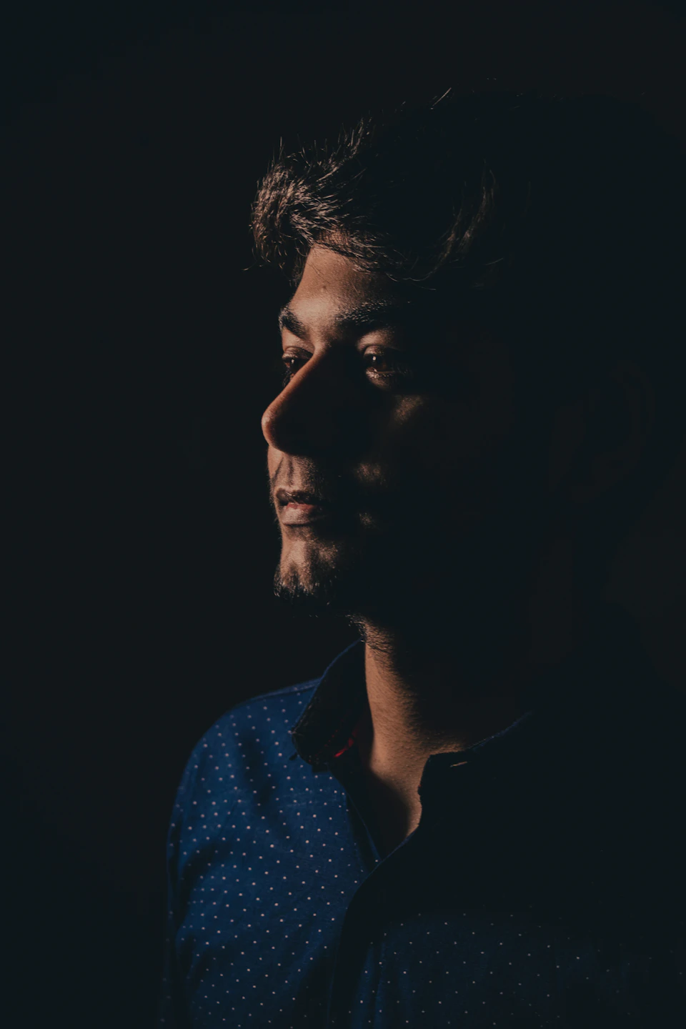 The image shows a man with short hair and a beard, wearing a blue shirt with white polka dots. He is looking off to the side, suggesting he might be deep in thought or lost in contemplation. The background is dark, which contrasts with his light skin tone and adds depth to the scene.