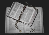 An open book with visible text is placed on top of another book. A rosary with beads and a cross is draped over the book, creating a religious and contemplative atmosphere.