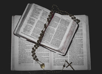An open book with visible text is placed on top of another book. A rosary with beads and a cross is draped over the book, creating a religious and contemplative atmosphere.