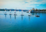A group of five sailboats with colorful numbered sails moves in a line across a calm body of water. A small motorboat with a few people on it follows alongside. In the background, industrial structures and trees line the shoreline under a blue sky with scattered clouds.