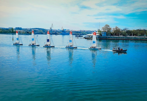 A group of five sailboats with colorful numbered sails moves in a line across a calm body of water. A small motorboat with a few people on it follows alongside. In the background, industrial structures and trees line the shoreline under a blue sky with scattered clouds.