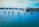 A group of five sailboats with colorful numbered sails moves in a line across a calm body of water. A small motorboat with a few people on it follows alongside. In the background, industrial structures and trees line the shoreline under a blue sky with scattered clouds.