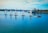 A group of five sailboats with colorful numbered sails moves in a line across a calm body of water. A small motorboat with a few people on it follows alongside. In the background, industrial structures and trees line the shoreline under a blue sky with scattered clouds.
