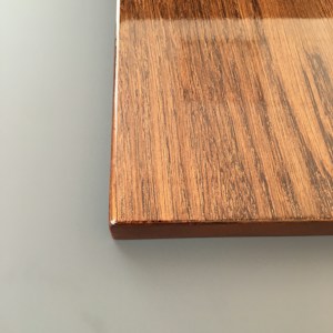 A close-up view of a rectangular wooden surface with a smooth finish. The wood grain is prominent and has a mixture of dark and light brown tones.