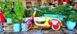 A colorful scooter is parked against a backdrop of lush, green plants in pots. The scooter is vibrantly painted with red, yellow, and turquoise, adorned with decorative flowers and a butterfly. The setting is outside a traditional-style building with a warm, rustic ambiance, featuring stone and wood elements.