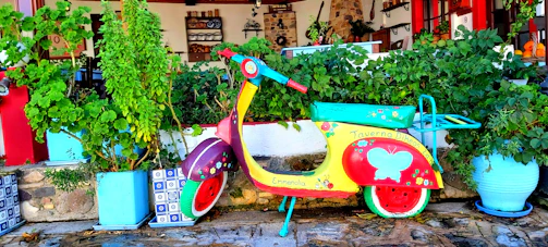A colorful scooter is parked against a backdrop of lush, green plants in pots. The scooter is vibrantly painted with red, yellow, and turquoise, adorned with decorative flowers and a butterfly. The setting is outside a traditional-style building with a warm, rustic ambiance, featuring stone and wood elements.