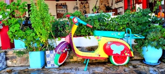A colorful scooter is parked against a backdrop of lush, green plants in pots. The scooter is vibrantly painted with red, yellow, and turquoise, adorned with decorative flowers and a butterfly. The setting is outside a traditional-style building with a warm, rustic ambiance, featuring stone and wood elements.