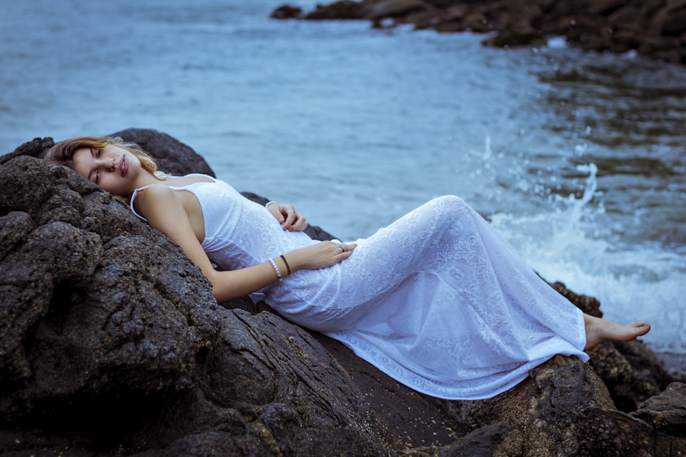 woman in white dress lying on rock near body of water during daytime