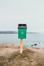 A green trash bin mounted on a wooden post is situated on a grassy area near a body of water, with a distant shoreline in the background under an overcast sky.