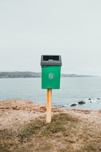 A green trash bin mounted on a wooden post is situated on a grassy area near a body of water, with a distant shoreline in the background under an overcast sky.