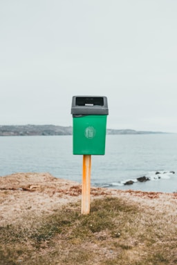 A green trash bin mounted on a wooden post is situated on a grassy area near a body of water, with a distant shoreline in the background under an overcast sky.