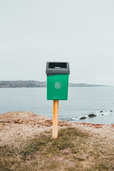 A green trash bin mounted on a wooden post is situated on a grassy area near a body of water, with a distant shoreline in the background under an overcast sky.