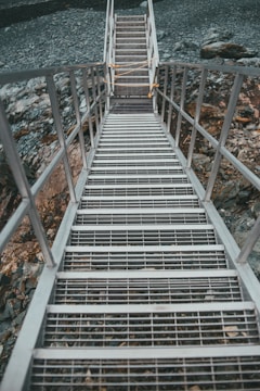 Metal stairs with grating steps lead downwards, surrounded by rocky terrain. A rope is tied across the upper section of the stairs, possibly as a safety measure.