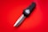A folding knife with a black handle and metallic blade is placed on a bright red surface. The blade is partially extended, showcasing its sharp edge and design details.