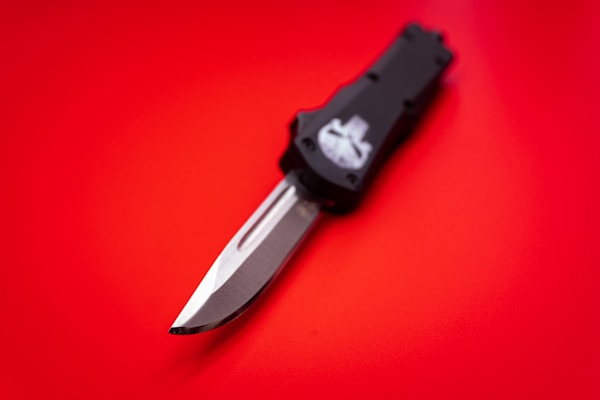 A folding knife with a black handle and metallic blade is placed on a bright red surface. The blade is partially extended, showcasing its sharp edge and design details.