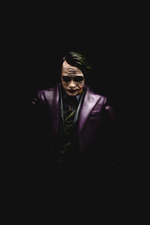 A dark and dramatic figure is centered against a black background, exhibiting pale white face paint with exaggerated dark eyes and a sinister expression. The character wears a dark purple jacket and a green shirt, with facial features highlighted by focused lighting from above.