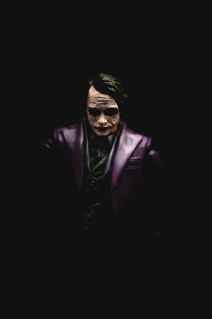 A dark and dramatic figure is centered against a black background, exhibiting pale white face paint with exaggerated dark eyes and a sinister expression. The character wears a dark purple jacket and a green shirt, with facial features highlighted by focused lighting from above.