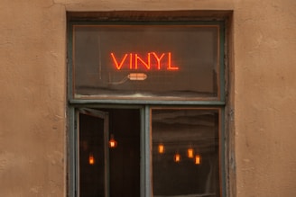 A window with a neon sign reading 'VINYL' in bright red letters. Below the sign, part of the window is open, revealing several warm colored pendant lights inside, creating a cozy and inviting atmosphere.
