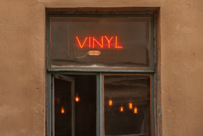 A window with a neon sign reading 'VINYL' in bright red letters. Below the sign, part of the window is open, revealing several warm colored pendant lights inside, creating a cozy and inviting atmosphere.