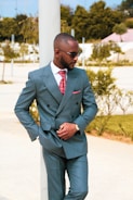 A man stands outdoors wearing a stylish blue double-breasted suit with a red patterned tie and pocket square. He is leaning casually against a column and wearing sunglasses, looking off to the side. The background features greenery and a park-like setting with trees and shrubs.