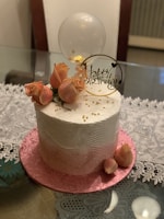 A beautifully decorated birthday cake with a smooth white frosting wave pattern. The cake is adorned with elegant pink roses and a clear balloon, along with a decorative sign that reads 'Happy Birthday'. Gold beads are scattered on the cake, adding a touch of luxury. The cake sits on a pink textured base, with a delicate lace tablecloth underneath.