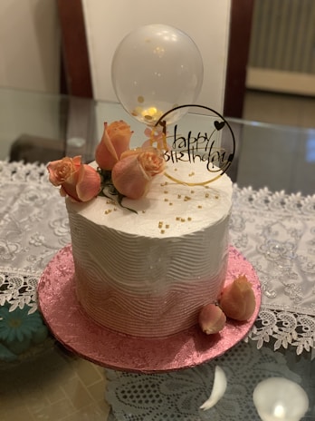 A beautifully decorated birthday cake with a smooth white frosting wave pattern. The cake is adorned with elegant pink roses and a clear balloon, along with a decorative sign that reads 'Happy Birthday'. Gold beads are scattered on the cake, adding a touch of luxury. The cake sits on a pink textured base, with a delicate lace tablecloth underneath.