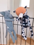 Two plush toys are hanging on a black metal railing. One toy resembles a blue elephant with a long nose, while the other is a spotted dog with an orange head. Both toys appear to be handmade and are positioned as if they are playfully hanging on the bars.