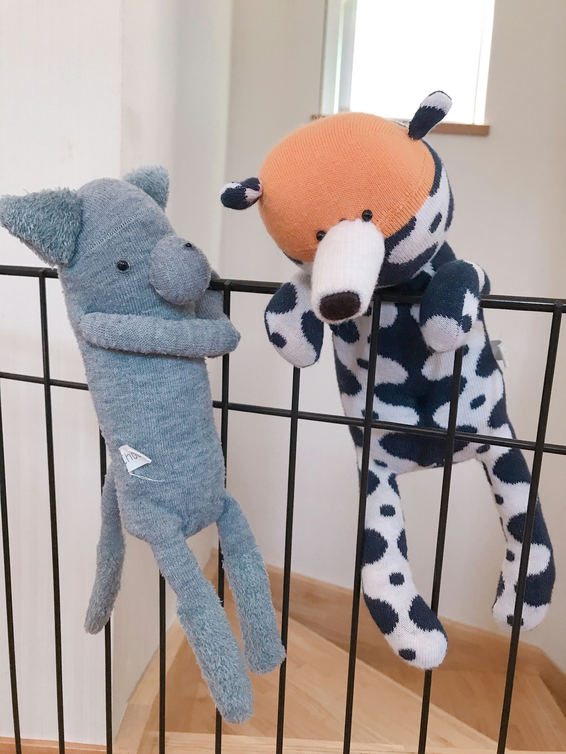 Two plush toys are hanging on a black metal railing. One toy resembles a blue elephant with a long nose, while the other is a spotted dog with an orange head. Both toys appear to be handmade and are positioned as if they are playfully hanging on the bars.