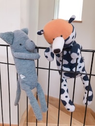 Two plush toys are hanging on a black metal railing. One toy resembles a blue elephant with a long nose, while the other is a spotted dog with an orange head. Both toys appear to be handmade and are positioned as if they are playfully hanging on the bars.