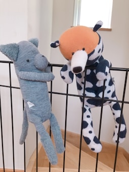 Two plush toys are hanging on a black metal railing. One toy resembles a blue elephant with a long nose, while the other is a spotted dog with an orange head. Both toys appear to be handmade and are positioned as if they are playfully hanging on the bars.