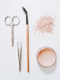 A flat lay arrangement of beauty tools and products on a white surface, including a pair of scissors, tweezers, a mascara wand with a wooden handle, a scattered amount of loose pink powder, and an open container filled with similar pink powder.
