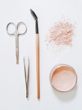 A flat lay arrangement of beauty tools and products on a white surface, including a pair of scissors, tweezers, a mascara wand with a wooden handle, a scattered amount of loose pink powder, and an open container filled with similar pink powder.