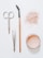 A flat lay arrangement of beauty tools and products on a white surface, including a pair of scissors, tweezers, a mascara wand with a wooden handle, a scattered amount of loose pink powder, and an open container filled with similar pink powder.