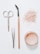 A flat lay arrangement of beauty tools and products on a white surface, including a pair of scissors, tweezers, a mascara wand with a wooden handle, a scattered amount of loose pink powder, and an open container filled with similar pink powder.