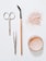 A flat lay arrangement of beauty tools and products on a white surface, including a pair of scissors, tweezers, a mascara wand with a wooden handle, a scattered amount of loose pink powder, and an open container filled with similar pink powder.