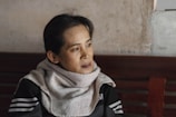 A person with short dark hair, draped in a light-colored scarf, sits contemplatively on a wooden bench. The background suggests a simple, possibly rustic indoor setting with a textured wall.