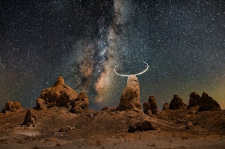 A stunning night sky filled with countless stars and the Milky Way galaxy vividly visible. Below, rocky formations rise from the ground, creating a surreal landscape. A crescent shape is seen around one of the rocky peaks, adding a mystical touch.