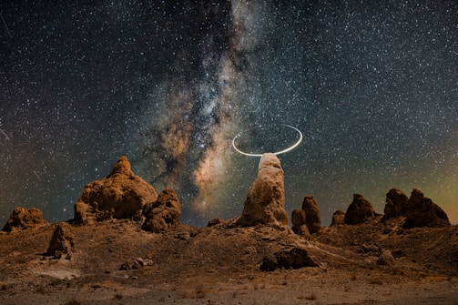 A stunning night sky filled with countless stars and the Milky Way galaxy vividly visible. Below, rocky formations rise from the ground, creating a surreal landscape. A crescent shape is seen around one of the rocky peaks, adding a mystical touch.