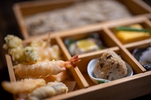 A bento box with various Japanese dishes, including tempura shrimp and vegetables, a portion of rice shaped into a ball with toppings, and several small side dishes, all neatly arranged in wooden compartments.