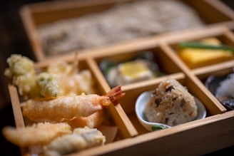 A bento box with various Japanese dishes, including tempura shrimp and vegetables, a portion of rice shaped into a ball with toppings, and several small side dishes, all neatly arranged in wooden compartments.