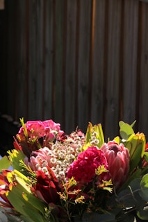 A vibrant bouquet of flowers with a mix of pink, red, and green hues under bright lighting. The flowers appear fresh and lush, with large blossoms surrounded by various leaves and smaller budding flowers. In the background, a wooden fence adds a rustic element to the composition.
