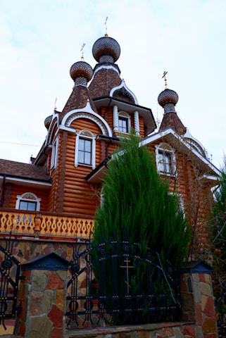 A traditional Russian-style wooden building featuring ornate onion domes with crosses sits behind an elaborate wrought iron fence. The structure is characterized by its rich brown log walls and white decorative trim. A large evergreen tree partially obscures the facade and adds a lush greenery to the scene.