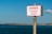 A warning sign mounted on a pole, positioned against a backdrop of a large body of water and a clear blue sky. The sign reads 'DANGER SLIPPERY SLOPE' in bold red letters on a white background, indicating a cautionary message.