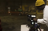 A man wearing a yellow beanie and a white shirt is operating a professional camera setup on a tripod, capturing footage of a brown, modified car performing a drift inside a warehouse with dim lighting. The warehouse has various signs and automotive equipment in the background.
