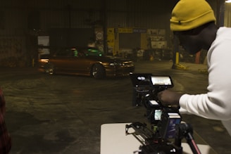 A man wearing a yellow beanie and a white shirt is operating a professional camera setup on a tripod, capturing footage of a brown, modified car performing a drift inside a warehouse with dim lighting. The warehouse has various signs and automotive equipment in the background.