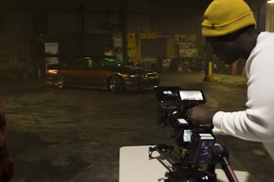 A man wearing a yellow beanie and a white shirt is operating a professional camera setup on a tripod, capturing footage of a brown, modified car performing a drift inside a warehouse with dim lighting. The warehouse has various signs and automotive equipment in the background.